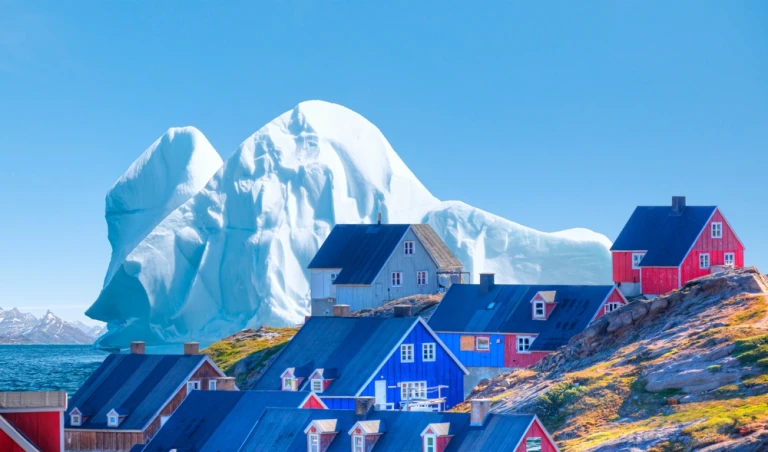 malerisches Dorf an der Küste Grönlands, ein riesiger Eisberg im Hintergrund - Farbige Häuser in Tasiilaq, Ostgrönland