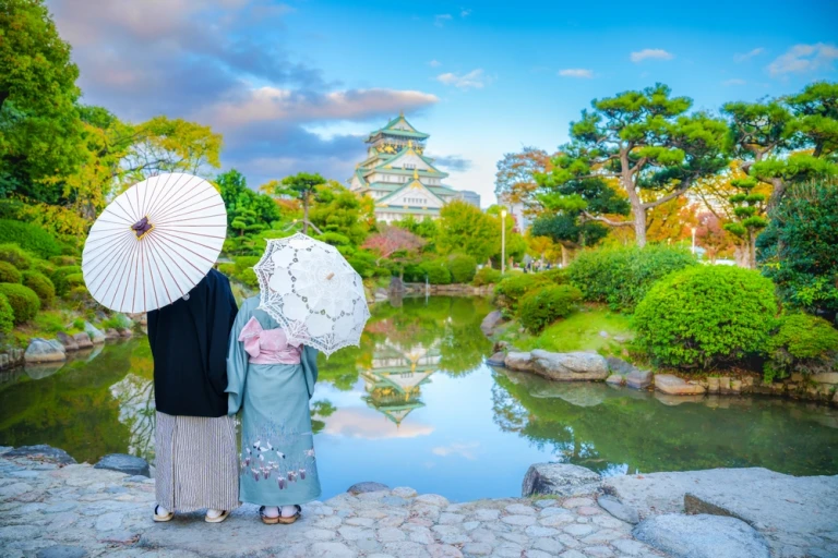 Ein asiatisches Liebespaar – ein Mann und eine Frau – in Kimonos, im traditionellen japanischen Stil, steht und blickt auf die Landschaft mit Blick auf die Burg Osaka bei Sonnenuntergang im Herbst, Osaka, Japan.